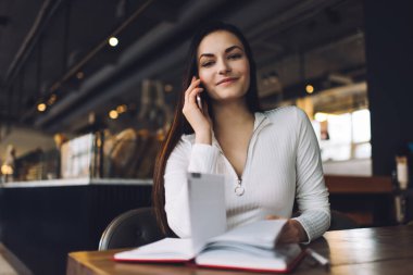 Neşeli genç bayan kameraya bakıyor ve gülümsüyor. Kafenin bulanık arka planında cep telefonundan önemli bir çağrı alıyor.