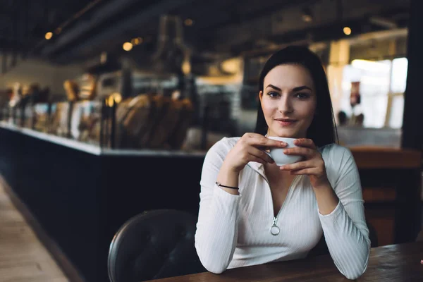 現代のカフェテリアで香り高いホットコーヒーを飲みながら 木製のテーブルに座っている間にカメラと笑顔を見て長い黒い髪を持つ魅力的な若い女性学生 — ストック写真