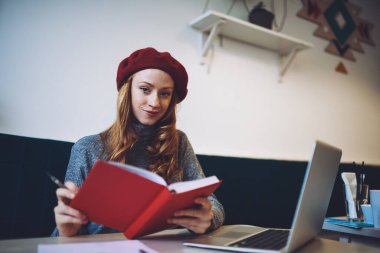 Bere takmış mutlu bir bayanın iş sırasında dizüstü bilgisayar ve not defteriyle masada kameraya bakmasından.