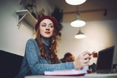 Kırmızı bereli mutlu bayanın kafeteryada çalışırken elinde dizüstü bilgisayarla masada oturmasından.