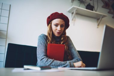 Kırmızı bereli gündelik kıyafetlerle gülümseyen ve iş sırasında masada elinde not defteriyle kameraya bakan bir genç.