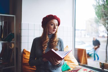 Düşünceli, uzun saçlı, mavi kazaklı ve kırmızı bereli bir kadın pencerenin yanındaki odada kitap okuyor ve başka tarafa bakıyor.