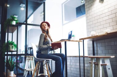 Moda elbiseli, kırmızı bereli, modern kafede dizüstü bilgisayarı ve dizüstü bilgisayarı olan genç bir kadın.