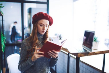 Moda kıyafetleri içinde genç bir bayan öğrenci test kitabı okuyor modern kafede dizüstü bilgisayarla masaya oturuyor.