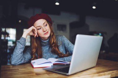 Genç pozitif kadın serbest çalışan, modern bir iş yerinde çalışırken dizüstü bilgisayar ve defterle masaya oturuyor.