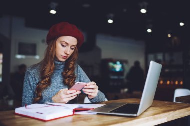 Konsantre kadın serbest çalışan, gündelik kıyafetli, dizüstü bilgisayarlı ve not defterli akıllı telefonlu, uzaktan kumandalı bir proje üzerinde çalışırken masada oturuyor.