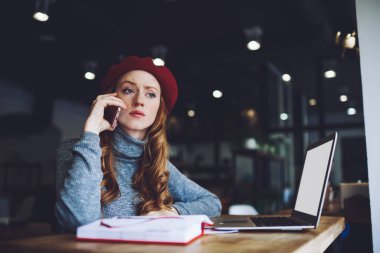 Bereli ve balıkçı yakalı, uzun saçlı, cep telefonundan arayan ve kafeteryada netbook üzerinde çalışan düşünceli bir kadın.