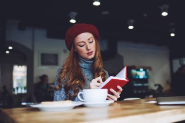 Kendine güvenen, boğazlı kazaklı, bereli, kırmızı kapaklı not defterine konsantre bir kadın modern kafede büyük bir fincan kahvenin tadını çıkarıyor.