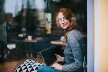 Genç, pozitif kadın, gözlüklü, sıradan giyinmiş, pencerenin kenarına bakan ve çalışırken bilgisayarını karıştıran serbest kadın.