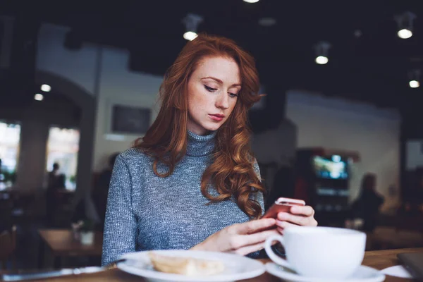 朝食中にモダンなカフェで時間を過ごしながら スマートフォンの画面に集中してカジュアルな服装で赤毛の女性の下から — ストック写真
