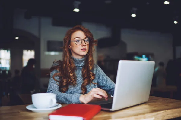 穿着休闲装 目光炯炯炯坐在桌旁 拿着咖啡和笔记本在笔记本电脑上工作的多愁善感的女性远程工作者 — 图库照片
