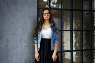 Kafkasyalı kot ceketli modanın yarı boy portresi şehir binasının yakınındaki kameraya bakıyor, optik gözlüklü beyaz kadın turist göz makyajı yapıyor.