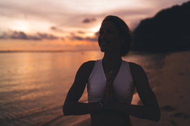 Okyanus kıyısında duran ve loş ışıkta tatil yaparken gün batımının tadını çıkaran genç pozitif kadın.