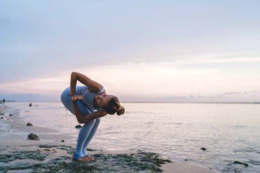 Aktif giyimli, çıplak ayaklı, genç bir kadın yaz mevsiminde dalgalı deniz yüzeyinde yoga yapıyor.
