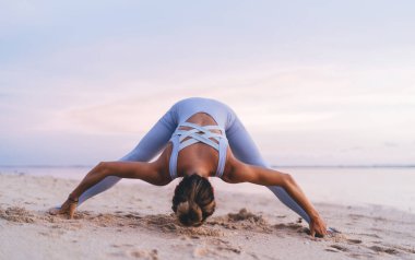 Aktif kıyafetli, çıplak ayaklı bir kadın yaz akşamları deniz suyunun yanındaki kumlu sahilde yoga yapıyor.