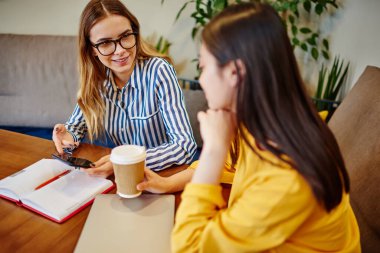 Şık giyinmiş neşeli çoklu etnik meslektaşlar elinde not defteriyle masada oturup kahve içip gündüz vakti kafede planlar hakkında konuşuyorlar.