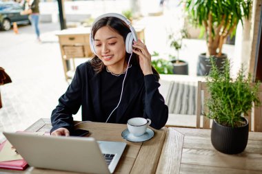 Geleneksel kıyafetli etnik bayan uzaktan çalışan cep telefonundan kulaklıkla müzik dinleyen ve verandada otururken içki ve not defterinin yanında çalışan.