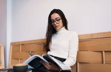 Beyaz süveter ve gözlüklü konsantre bir kadın modern kafede sıcak kahve içerken rahatlıyor.