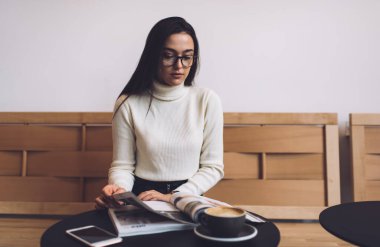 Normal kıyafetler ve gözlüklerle sakin bir kadın. Yuvarlak ahşap masada oturuyor ve modern kafede dinlenirken dergi okuyor.