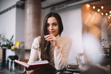 Gülümseyen kadın, kafede kahve molasında masada otururken kitap okuyor ve elinde gözlük tutuyordu.