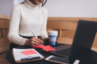 Ürün odaklı kadın girişimci günlük kıyafet notları yazarken ve içerken restoranda tek başına dizüstü bilgisayar ve not panosuyla otururken.
