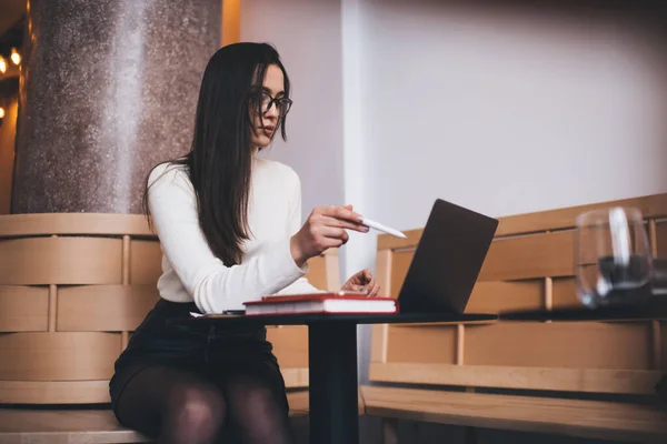 stock image Anonymous woman in elegant white turtleneck and black skirt sitting at round table with notebook and browsing laptop in cozy workplace