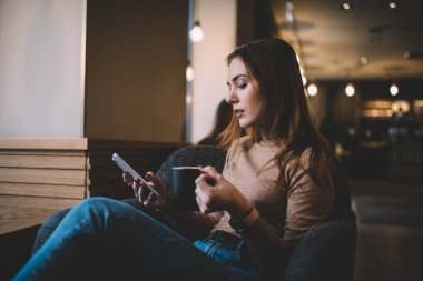 Odaklanmış bir kadının cep telefonuna bakması ve sıcak kahve içmesi koltukta tek başına oturması ve buluşmayı beklemesi.