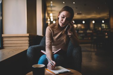 Konsantre olmuş genç bir kadın, masada pano ile oturuyor ve akıllı telefonla konuşurken notlar alıyor ve uzaktan bağımsız olarak çalışıyor.