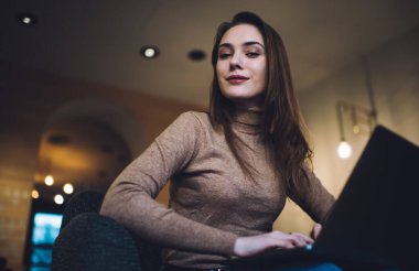 Günümüz kafeteryasında dizüstü bilgisayar kullanarak tek başına çalışan ve kameraya bakan kendine güvenen düşük açılı bir kadın.