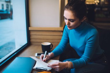 Pencerenin yanında oturan ve rahat çalışma alanında proje üzerinde çalışırken panoya notlar yazan ciddi bir kadının yüksek açısı