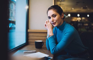 Mavi boğazlı, zarif kol saati olan genç bir kadın. Kafe masasında elinde not defteri ve kapalı dizüstü bilgisayarla kameraya bakıyor.