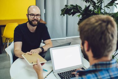 Optik gözlüklü sakallı erkek bilişim uzmanı. İnternet siteniz için taklit alanı olan dizüstü bilgisayarı kullanan yetenekli meslektaşınıza eğitim kitabı veriyor. Grafik tasarımcılar işbirliği yapıyor.