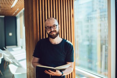 Eğitimli, ders kitabı öğrenen ve üniversitede okuyan zeki bir erkek öğrenci, elinde fotokopi kitabı tutarak göz düzeltme sağlayan optik gözlüklü, beyaz tenli hippi portresi.
