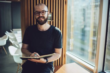 Eğitimli, ders kitabı öğrenen ve üniversitede okuyan zeki bir erkek öğrenci, elinde fotokopi kitabı tutarak göz düzeltme sağlayan optik gözlüklü, beyaz tenli hippi portresi.
