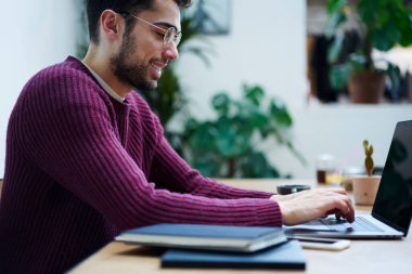 Modern kafede oturan ve netbook 'ta yazan, günlük kıyafetleri ve gözlükleriyle gülümseyen etnik sakallı bir erkek.
