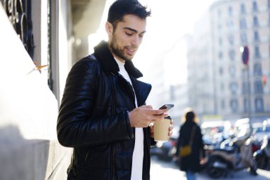 Ciddi genç sakallı erkek sıcak bir ceket giyiyor cep telefonuna bakıyor ve elinde bir fincan kahve tutarak sokakta vakit geçiriyor.