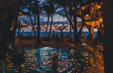 Bali 'de tatilde, karanlıkta aydınlık lambalarla çevrili, havuzda yüzen, tanınmayan bir kadının yüksek açısı.