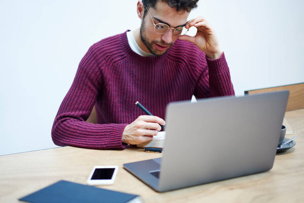 Бородатый этнический фрилансер в повседневной одежде и очках сидит за деревянным столом с ноутбуком и пишет идеи в блокноте