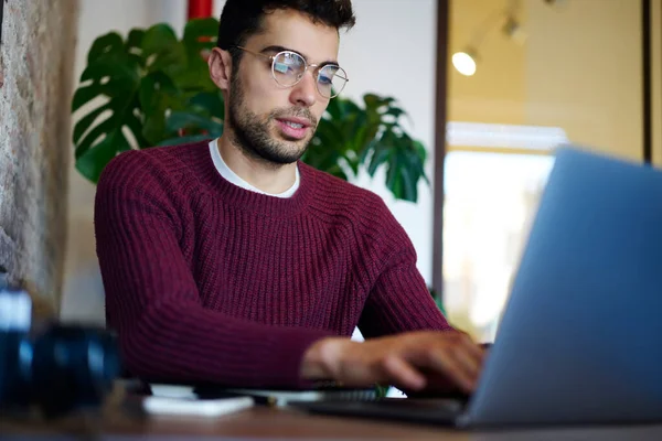 穿着紫色毛衣和眼镜的专心致志的男性自由职业者在用植物装饰的咖啡店工作时 用笔记本电脑打字 — 图库照片