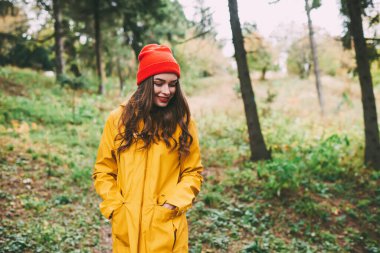 Sarı yağmurluklu ve şapkalı bir kadın elleri ceplerinde ormanda boş boş boş otururken aşağıya bakıyor.