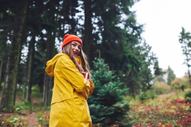 Kalın elbiseli genç bir kadının yeşil ormanda elinde kozalaklı ağaçlarla kameraya baktığı alçak açı.