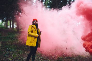 Parlak yağmurluklu ve şapkalı düşünceli genç bir kadın ormanda tek başına yürürken pembe duman bombasının yanında duruyor.