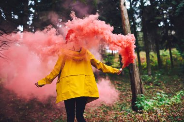 Sarı yağmurluklu, elleri ayrık duran, ormandaki duman bombasının altında kırmızı dumanlı, tanınmayan bir kadının arkası.