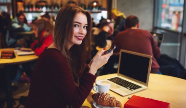 Günlük giysiler içinde gülümseyen bir kadın kameraya bakarken masada kruvasan ve bir fincan kahve içen akıllı telefonuyla oturuyor.