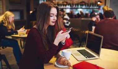 Açık dizüstü bilgisayar ve bir fincan lezzetli kahveyle masada oturan serbest çalışan mutlu kadın akıllı telefonlara göz gezdirirken...
