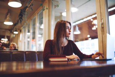 Düşük açılı, gündelik giysiler içinde, pencerenin yanındaki masada oturmuş çalışırken dizüstü bilgisayarda yazan kadın uzaktan kumanda.