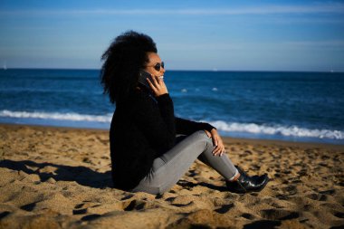 Tüm vücudun yandan görünüşü, günlük kıyafet ve güneş gözlüğü takan mutlu Afro-Amerikan kadın kumlu sahilde oturuyor ve akıllı telefondan arıyor.