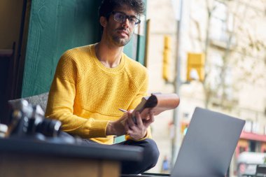 Sıradan kıyafetler ve gözlüklerle dizüstü bilgisayarla oturup not defterine notlar yazan düşünceli etnik erkek serbest yazar.