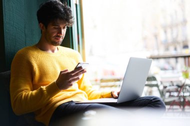 Rahat bir kafede rahat bir şekilde dinlenen günlük giysiler içinde genç etnik erkek çalışan ve uzaktan bilgisayarla çalışırken sosyal medyayı karıştırıyor.