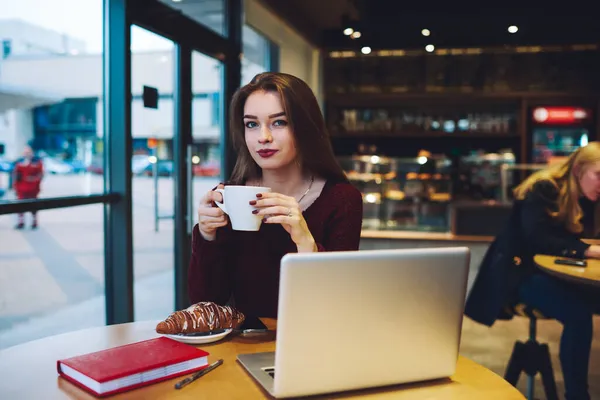 여성은 커피를 마시고 일하고 컴퓨터와 탁자에 카메라를 기뻐하고 — 스톡 사진